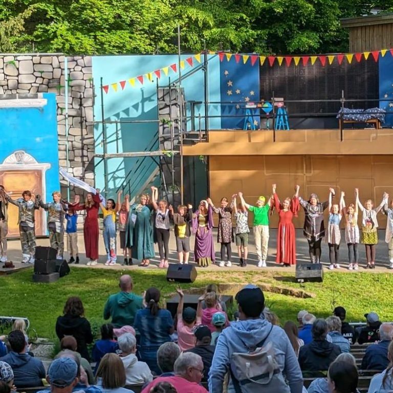 Musicalaufführung im Freien mit Darstellern auf der Bühne und Publikum im Vordergrund.