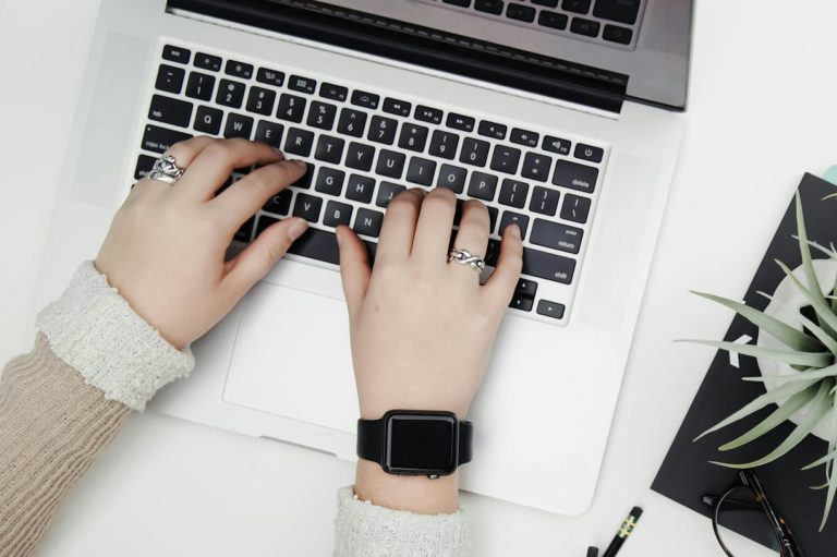 Hände tippen auf einer Laptop-Tastatur, neben einer Pflanze und einem Notizbuch.