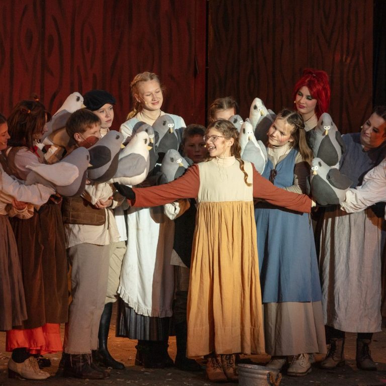 Eine Gruppe von Menschen in historischen Kostümen umarmt eine zentrale Figur mit Tauben.