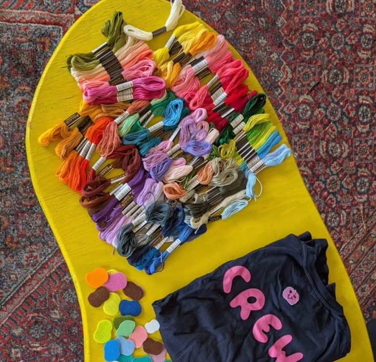 Bunte Wollfäden und ein schwarzes T-Shirt mit dem Text "CREW" auf einem gelben Untergrund.