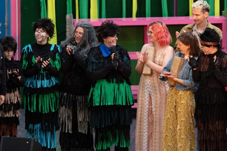 Eine Gruppe von Personen in bunten Kostümen steht applaudierend auf einer Bühne.