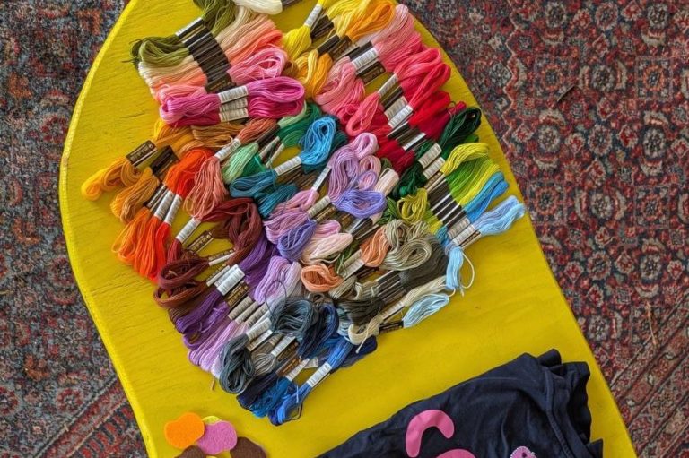 Bunte Garne in verschiedenen Farben auf einem gelben Untergrund, darunter ein dunkelblaues Shirt.