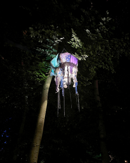Farbige Laterne in Form einer Qualle hängt in einem Baum, umgeben von dunklem Wald.